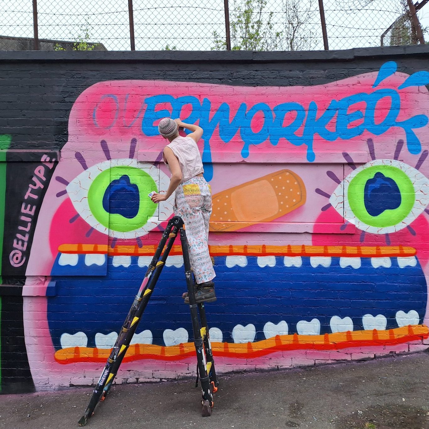 Künstler malt "Overworked" in Neonfarben beim Glasgow Graffiti Festival Street-Art-Künstler ellitype malt leuchtend-blaues E auf Graffiti-Schriftzug "Overworked", Rückenansicht beim Glasgow Graffiti Festival