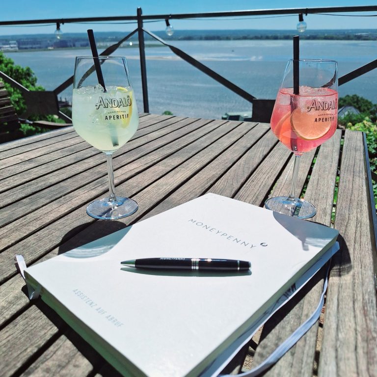 Ein Notizbuch und ein Kugelschreiber mit 2 Cocktails bei strahlend blauem Himmel auf der Terrasse eines Restaurants mit Elbblick